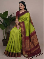 Load image into Gallery viewer, Woman wearing a green saree with maroon and gold border, posing in front of plants.