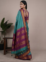 Load image into Gallery viewer, Woman wearing a turquoise saree with a purple blouse, posing in front of plants.