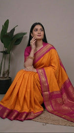 Load image into Gallery viewer, Woman wearing an orange saree with a pink border, sitting and posing against a neutral background.