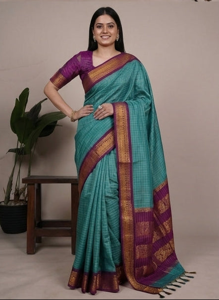 Woman wearing a turquoise saree with a purple blouse, posing in front of plants.