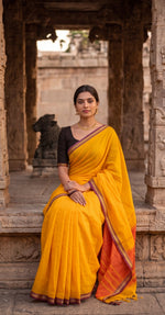 Load image into Gallery viewer, Woman in a yellow saree with an orange border, posing in front of stone architecture.