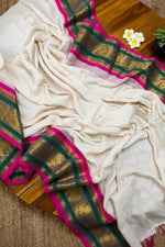 Load image into Gallery viewer, An off-white silk saree with a wide gold, green, and bright pink border, draped elegantly on a wooden surface with a small succulent and a brand card.