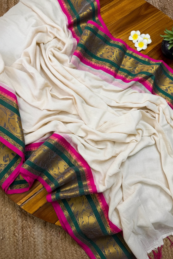 An off-white silk saree with a wide gold, green, and bright pink border, draped elegantly on a wooden surface with a small succulent and a brand card.